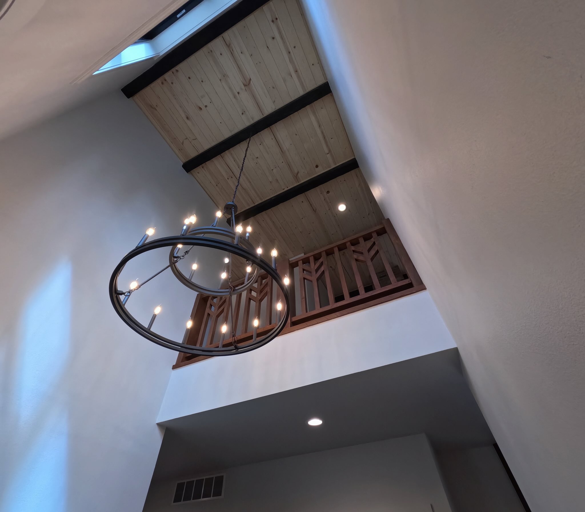 Looking straight up a two-story entry at a ring chandelier with candle bulbs, a pine tongue-and-groove ceiling with dark beams, a skylight and a wood loft railing with chevron-pattern balusters